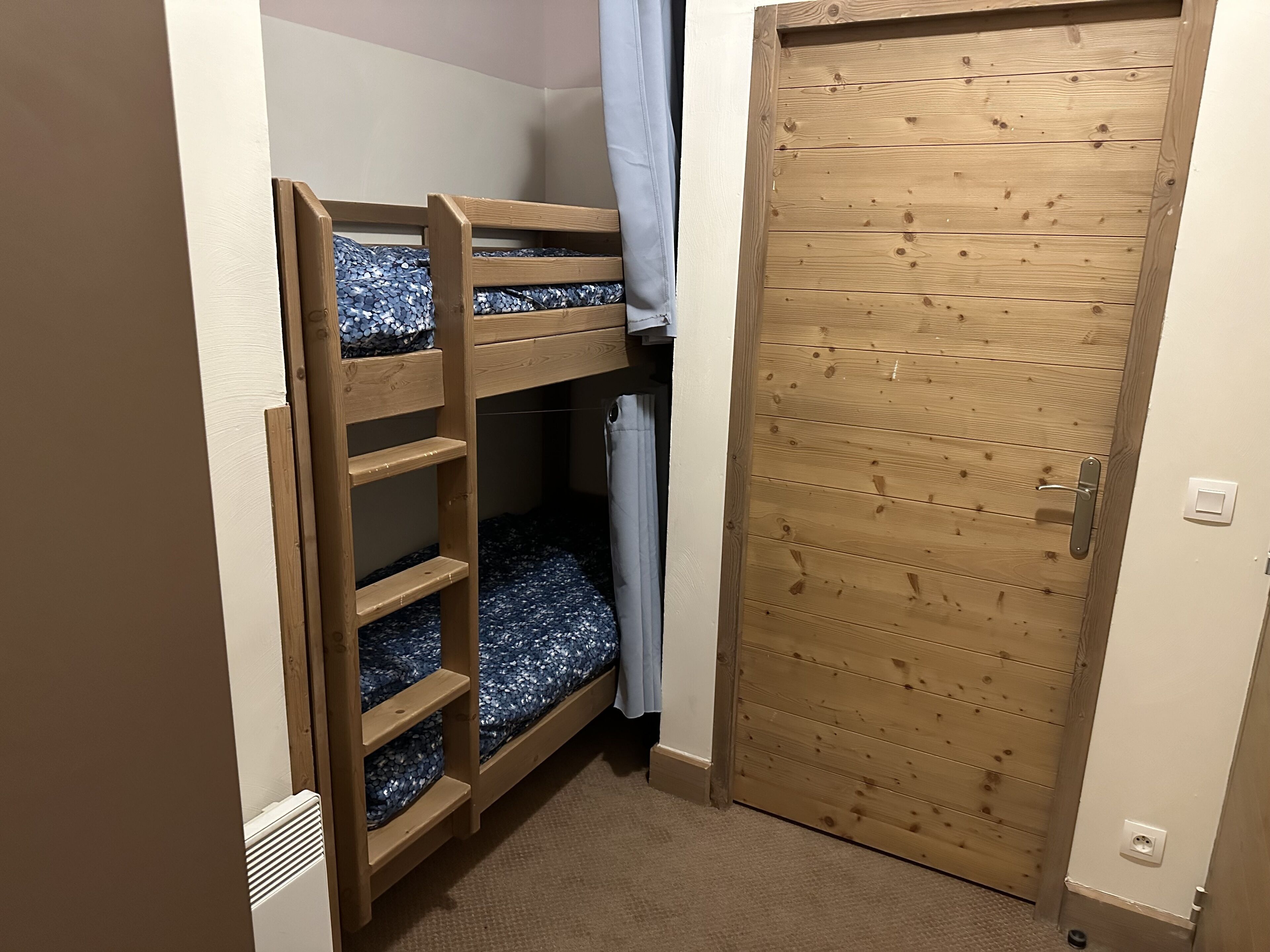 A bunk bed with a wooden frame and a blue patterned mattress, a wooden door with a handle, and a light switch on the wall.