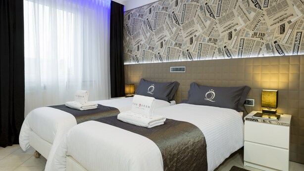 Habitación de hotel con dos camas, un ventanal con cortinas translúcidas, una pared con estampado de periódico y una mesita de noche con lámpara.