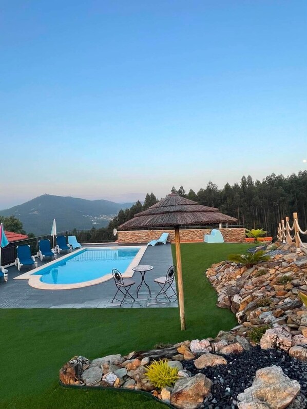 A swimming pool with lounge chairs and a thatched umbrella, surrounded by a landscaped garden with a view of distant mountains.