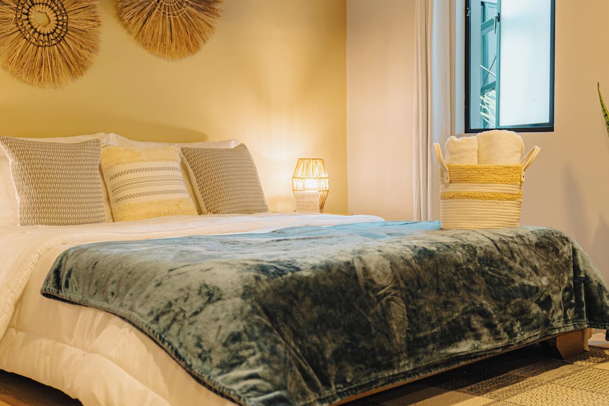 A bedroom with a bed, a window, a wall-mounted air conditioner, and a basket of towels.