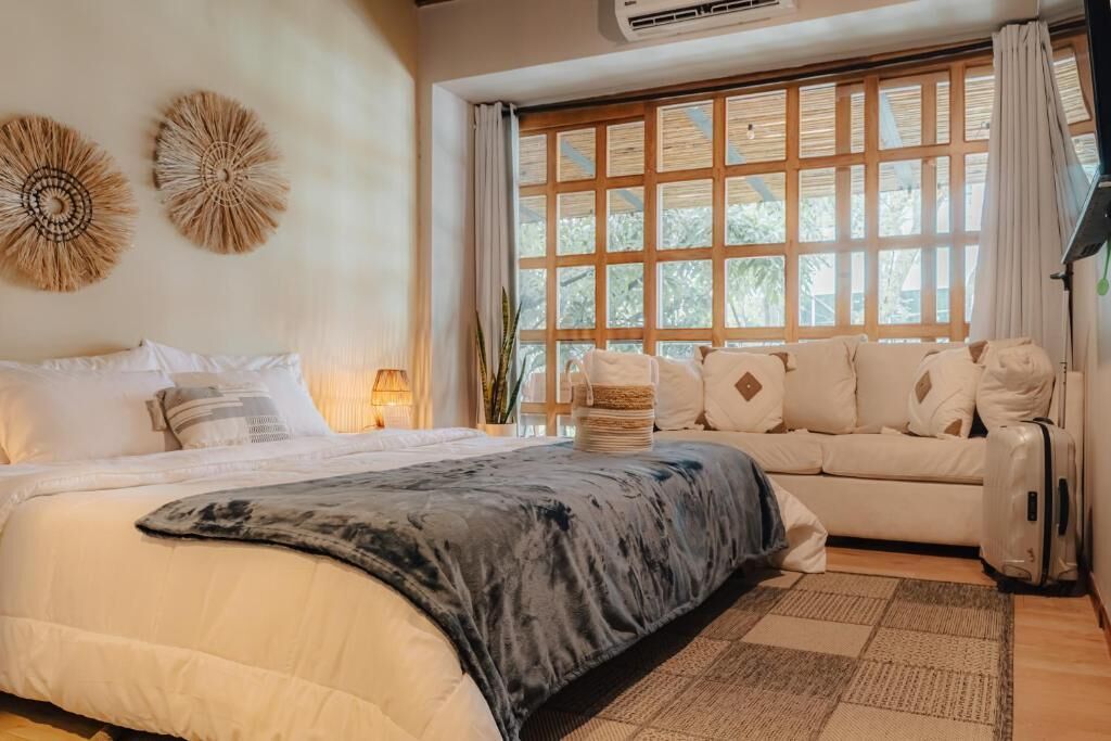 A bedroom with a bed, a sofa, a window with blinds, and a wall-mounted air conditioner.
