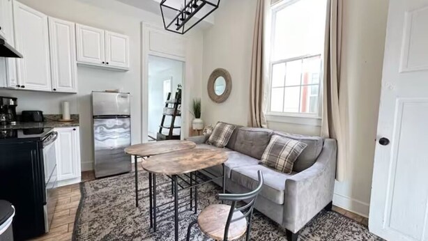 Una cocina moderna con armarios blancos, encimera de mármol y frigorífico empotrado. Hay una zona de comedor con un sofá gris y una mesa redonda.