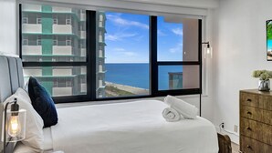 Hotelkamer met een groot raam dat uitzicht biedt op de oceaan en gebouwen. Er staat een bed met witte lakens, een houten dressoir en een bedlampje.