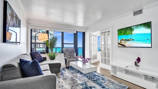 Een moderne woonkamer met een grijze bank, een fauteuil met een patroon en een salontafel met paarse bloemen. Een grote tv aan de muur toont een strandbeeld.