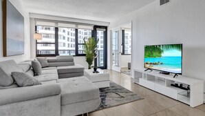 A modern living room with a grey sectional sofa, a flat-screen TV, and a large window overlooking a cityscape. - Cozy 2 Bedroom Ocean View / Beach Service / 707 (Miami Beach)
