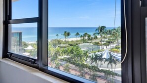A view from a window overlooking a beach resort with palm trees, a pool, and a clear blue sky. - Cozy 2 Bedroom Ocean View / Beach Service / 707 (Miami Beach)