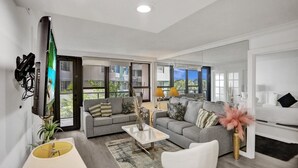Ein modernes Wohnzimmer mit grauem Sofa, weißem Sessel und Couchtisch. Ein Fernseher ist an der Wand montiert und ein Fenster bietet Blick auf Gebäude.