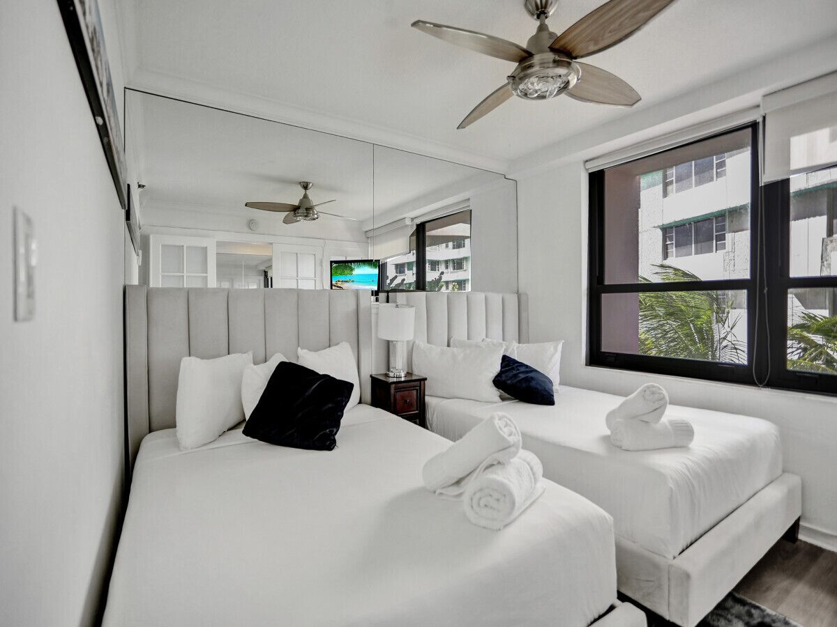 A hotel room with two beds, a ceiling fan, a TV, and a window with a view of buildings and greenery.