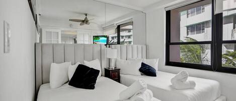 A hotel room with two beds, a ceiling fan, a TV, and a window with a view of buildings and greenery.