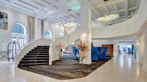A grand lobby with a central staircase, a chandelier, and a reception area with blue seating. - Cozy Ocean View Condo / Beach Service & Pool /808 (Miami Beach)