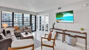 A modern living room with a large TV, comfortable seating, and a view of the city through large windows. - High Floor Beachfront Condo / Beach Service / 1509 (Miami Beach)
