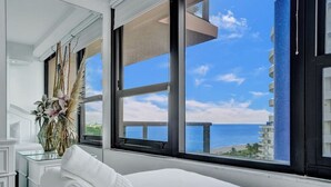 Een hotelkamer met een groot bed, een glazen tafel met een bloevas en uitzicht op de oceaan door grote ramen.