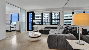 Una sala de estar moderna con un sofá gris, una mesa de centro redonda y una lámpara de pie. Amplias ventanas ofrecen vista a la ciudad.