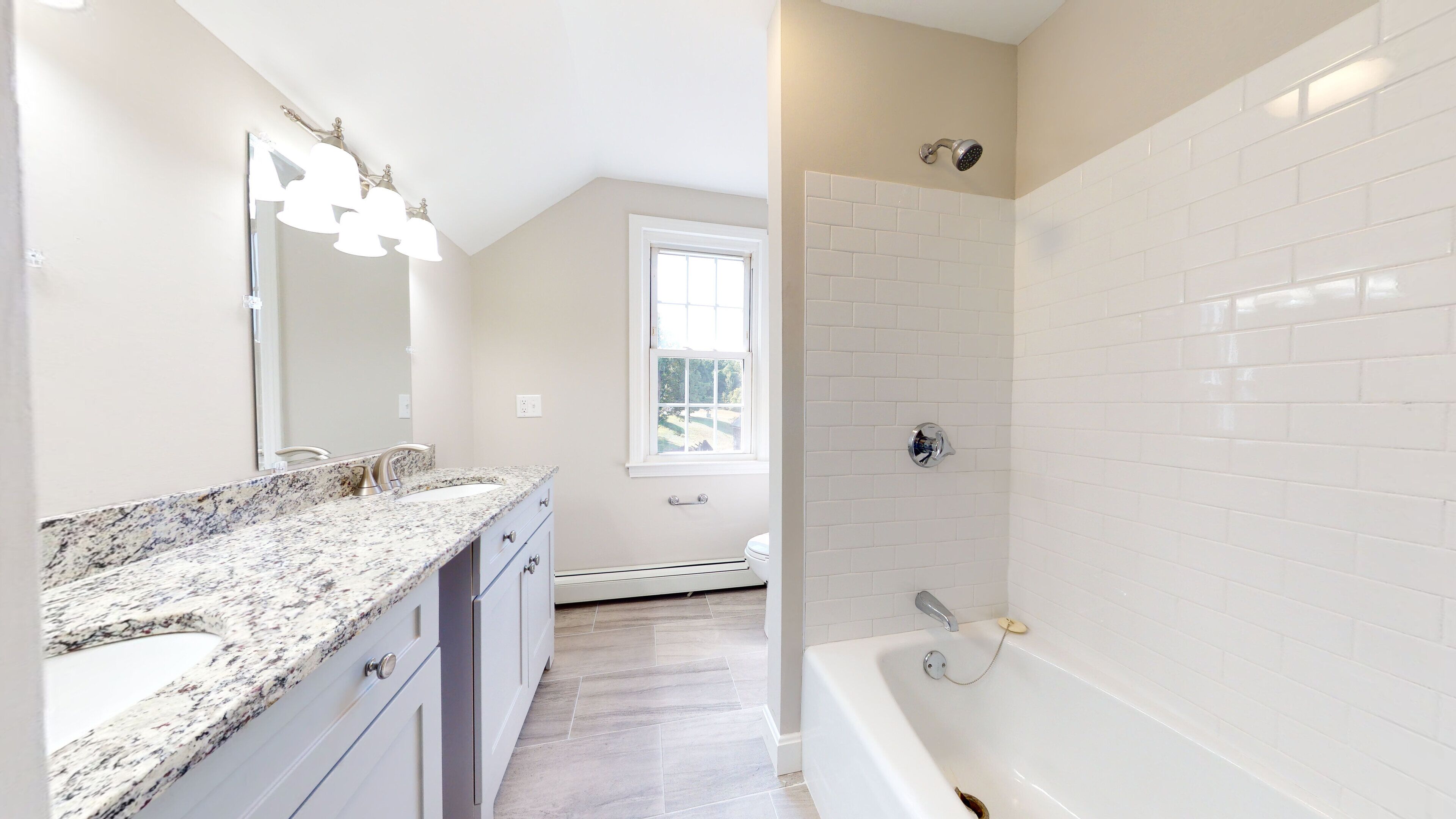 A modern bathroom with a granite countertop, a large mirror, a shower with a handheld showerhead, and a window providing natural light.