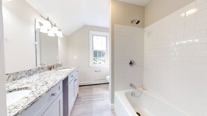 A modern bathroom with a granite countertop, a large mirror, a shower with a handheld showerhead, and a window providing natural light. - Central, Charming & Modern Home (Lakeville)