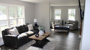 Ein Wohnzimmer mit einem schwarzen Ledersofa, einem Holztisch und einem beige farbenen Sessel. Zwei Fenster mit Jalousien, ein flachbildfernseher an der Wand und ein großes Fenster mit Blick auf Grünfläche.