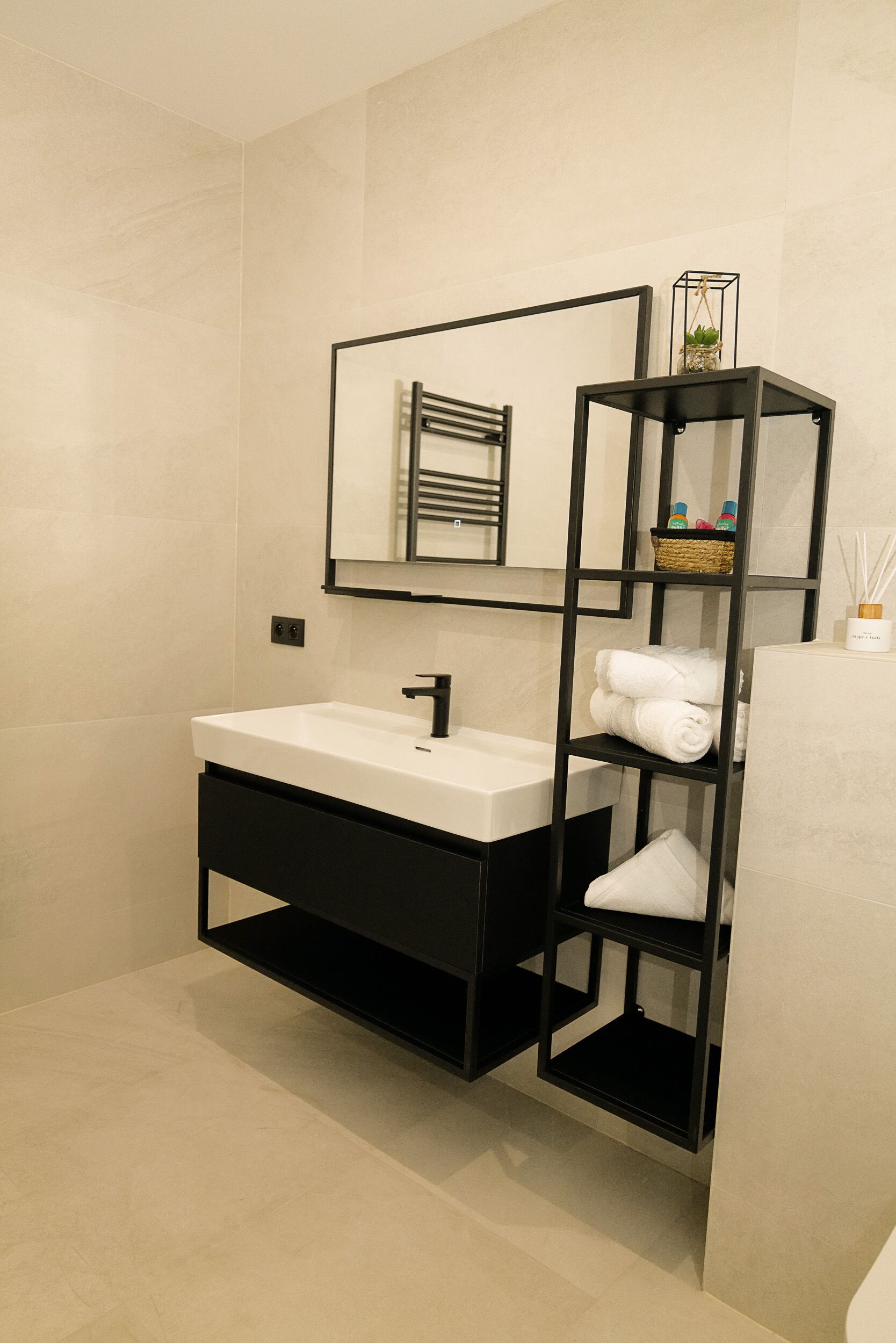 Un baño moderno con un lavamanos blanco, estanterías negras y una pared espejada.