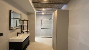Une salle de bain moderne avec un lavabo blanc, un meuble-lavabo noir et un grand miroir. Le plafond est doté de briques apparentes et d’un éclairage encastré.