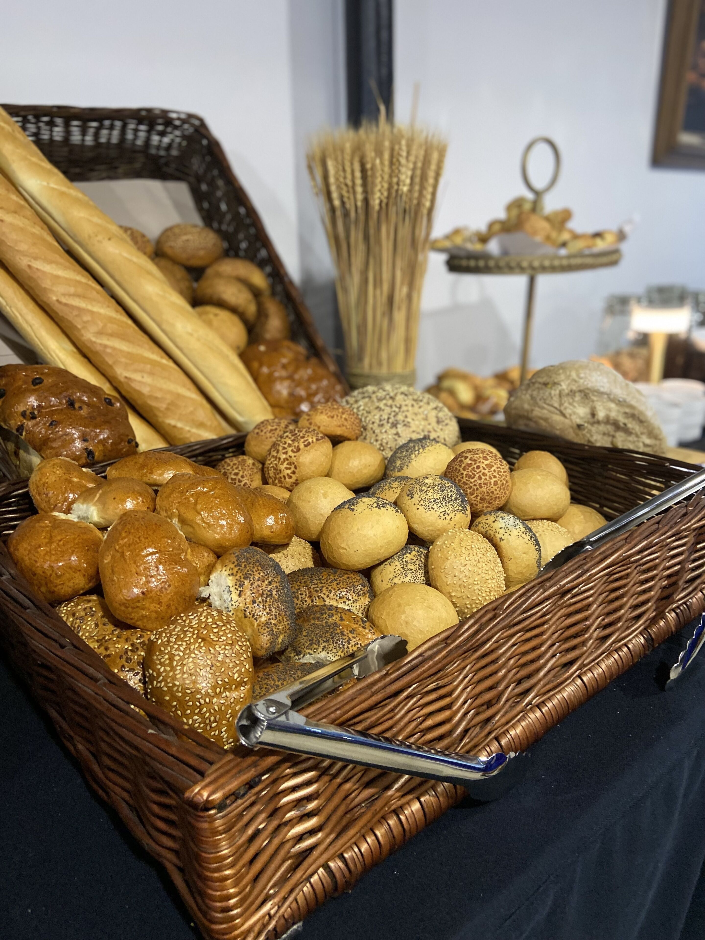 A wicker basket filled with various types of bread and rolls, including baguettes, rolls, and sesame seed buns.