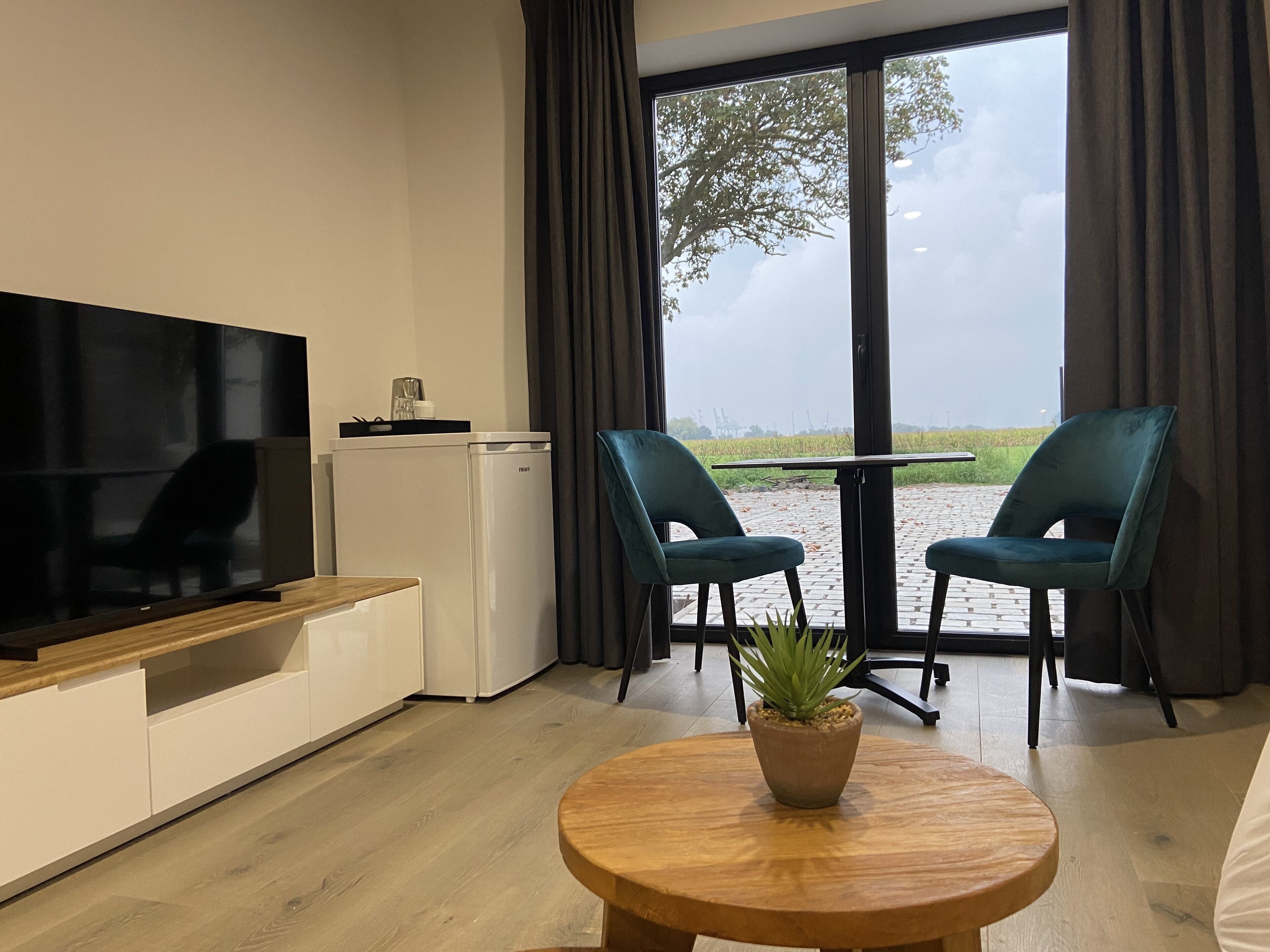Un saló modern amb televisió de pantalla plana, nevera petita, dues cadires, taula rodona de fusta amb una planta en test i una finestra gran amb vista a un camp i edificis allunyats.
