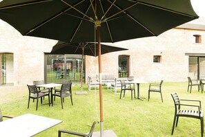 Un patio al aire libre con mesas y sillas, un paraguas grande y un edificio de ladrillo al fondo.