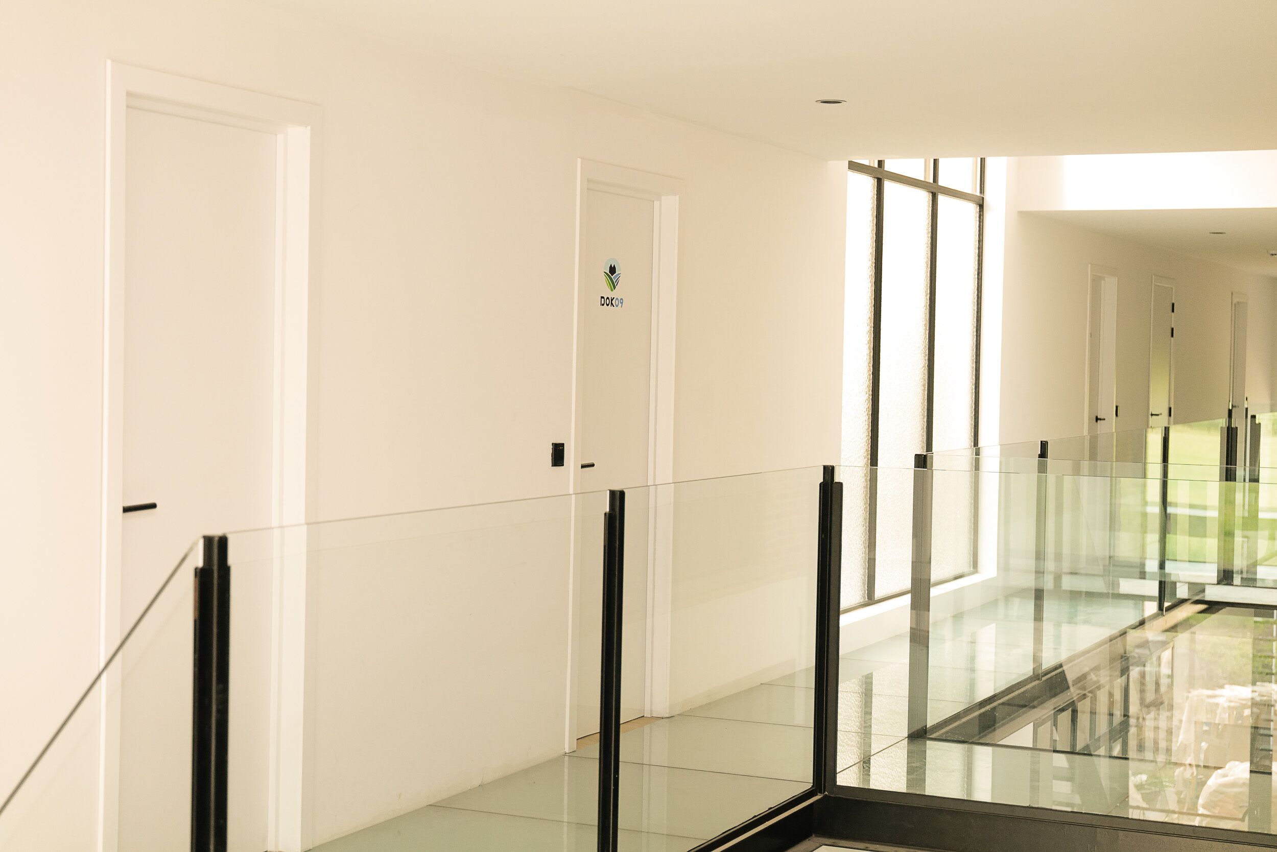 A modern hotel corridor with glass railings, white walls, and a visible door with a logo.