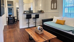 Una cocina moderna con gabinetes blancos, un microondas y una pequeña isla. Hay una zona de comedor con una mesa de madera y dos taburetes negros. En la sala de estar hay un sofá de cuero negro con una manta azul y una almohada mostaza.
