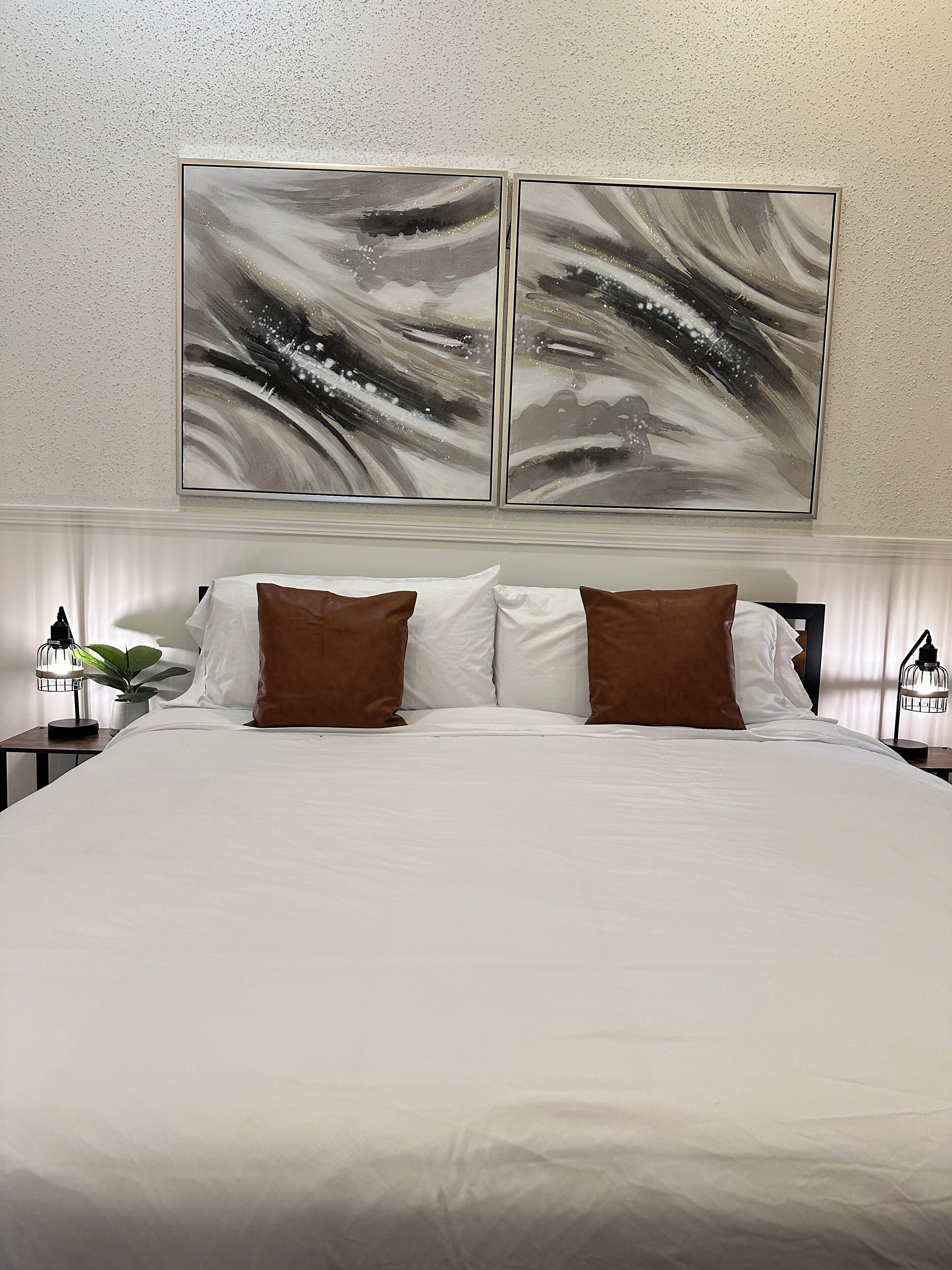 Una cama grande con ropa de cama blanca y marrón, dos mesitas de noche con lámparas, y un cuadro abstracto colgado en la pared.