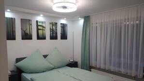 Ein Schlafzimmer mit Bett, Nachttisch, Heizkörper und einem Fenster mit durchsichtigen Vorhängen.