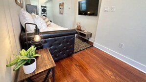 Une chambre à coucher moderne avec un lit, une table de chevet avec une lampe, une plante en pot et un téléviseur fixé au mur.