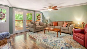 A living room with two sofas, a coffee table, and a red armchair. There is a ceiling fan and a large window with a view of greenery. - Cameo Tree Top Chalet - HOT TUB and Pet Friendly (Jasper)