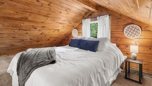 Un dormitorio acogedor con techo de madera, una cama con ropa de cama blanca y azul, un plaid gris y una mesita de noche pequeña con una lámpara.