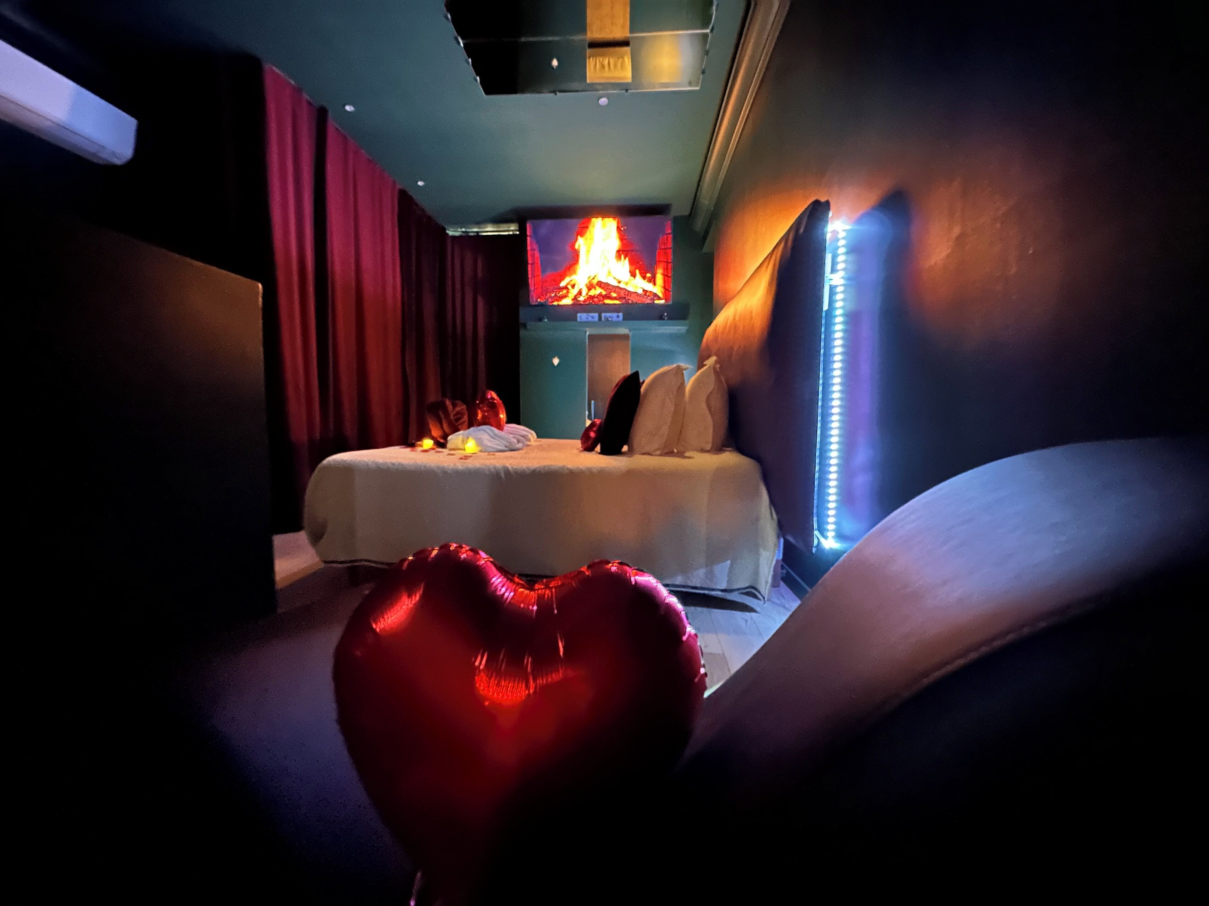 Una habitación de hotel con cama, almohada con forma de corazón y televisor de pared mostrando un fuego.