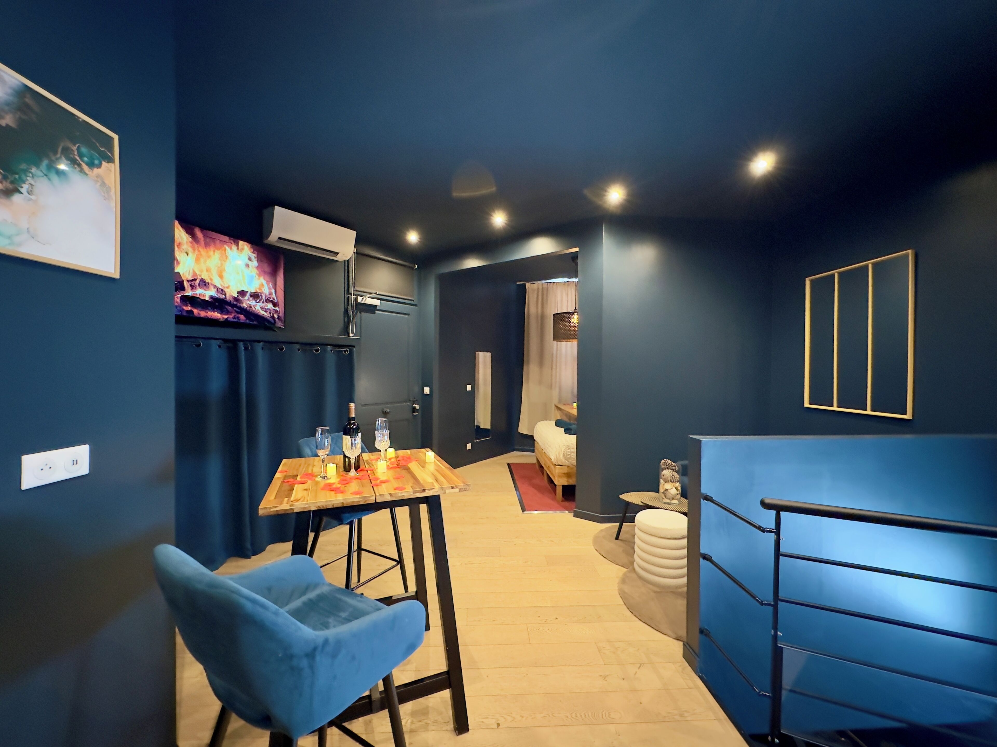 Una habitación moderna con una pared azul oscuro, una mesa de madera con velas, una silla azul y una cama al fondo.