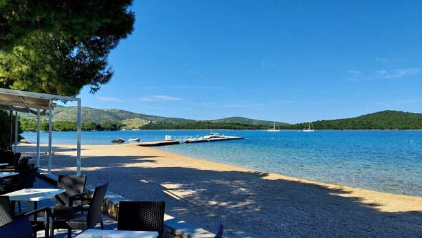 A beachside restaurant with outdoor seating, a dock with boats, and a view of a lake with hills in the background. - Apartment in Žaborić with Seaview, Balcony, Air condition, WIFI (5177-2) (Šibenik)