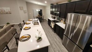 A modern kitchen with a long island, dark cabinets, stainless steel appliances, and a dining area with chairs and a table.