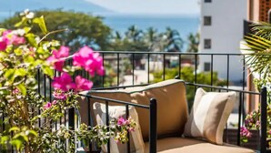 A balcony with a beige sofa, pink flowers, and a view of the sea and buildings. - 2BR-2BA Heart of Romantic Zone + Infinity pool (Puerto Vallarta)