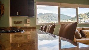 A kitchen with a large island, a dining area with chairs, and a view of mountains through a window. - 2BR-2BA Heart of Romantic Zone + Infinity pool (Puerto Vallarta)