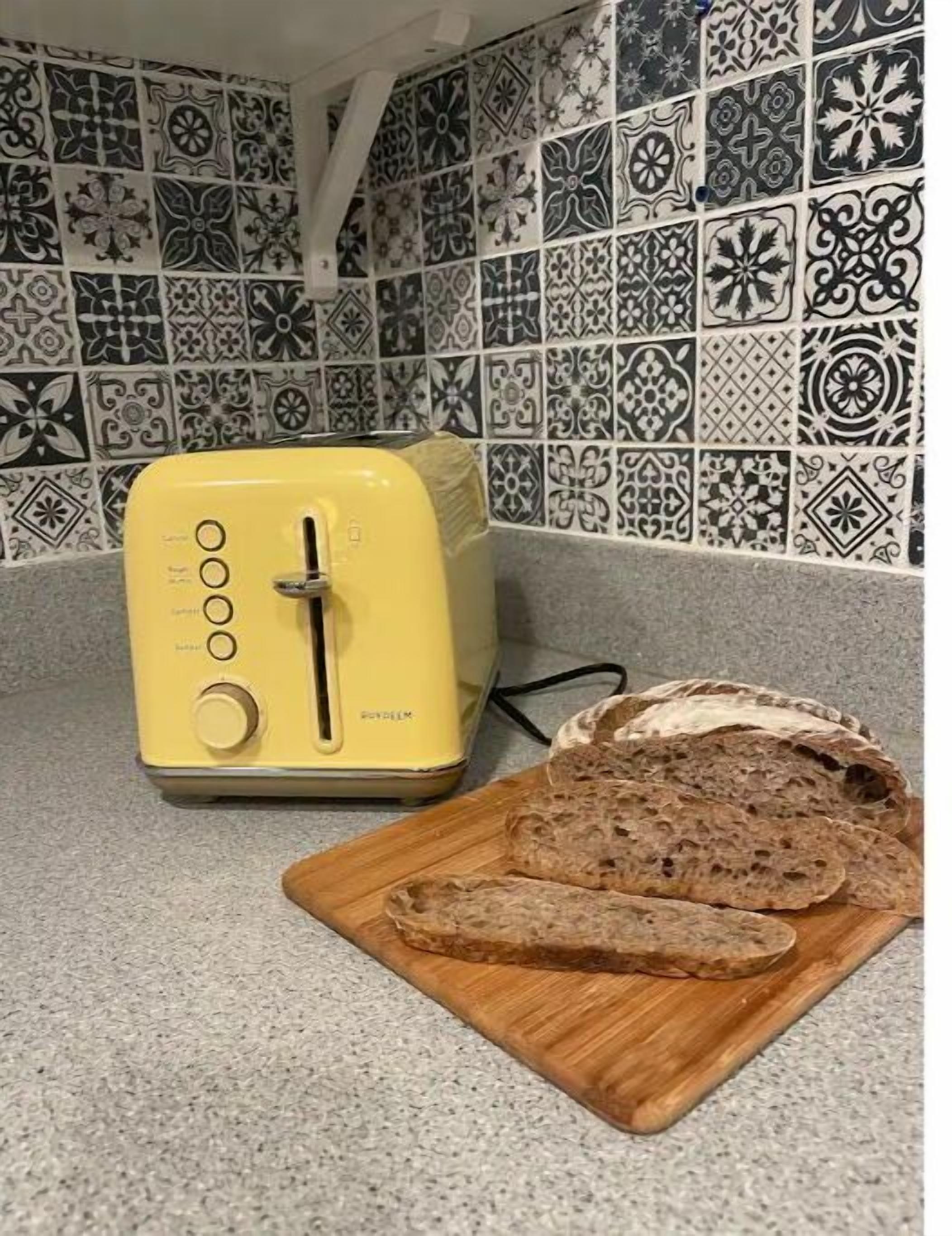Un tostador amarillo sobre una encimera de cocina con un salpicadero estampado y una tabla de cortar de madera con pan rebanado.