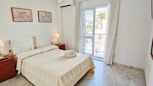 Un dormitorio con cama, dos mesitas de noche con lámparas, televisión y balcón con vistas a edificios y palmeras.