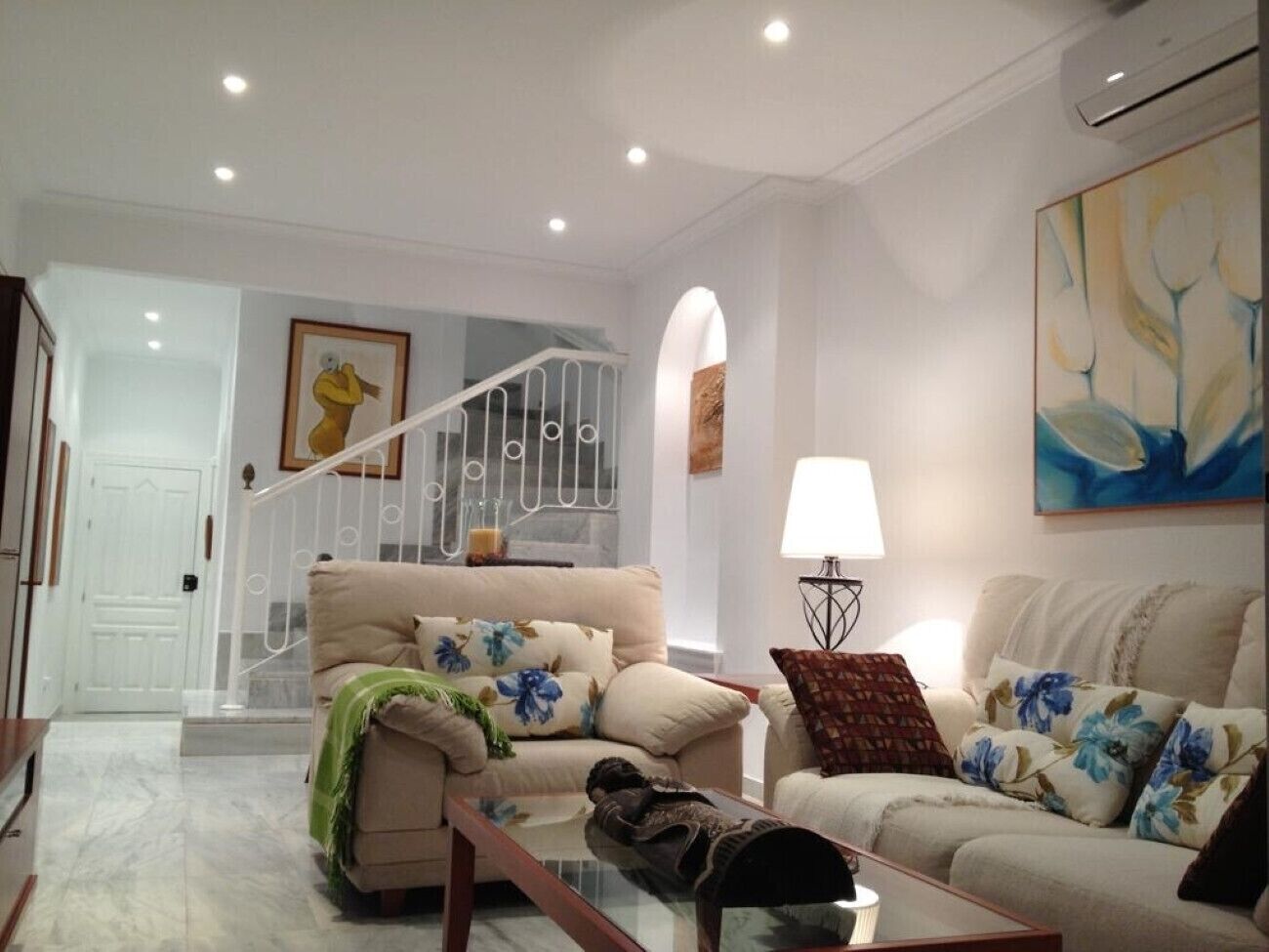 A spacious living room with a beige sofa, a green throw blanket, and a coffee table. A staircase with a white railing leads to another level.