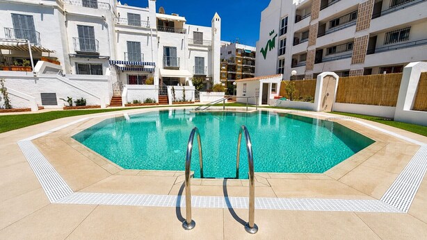 Una piscina con dos escaleras de metal, rodeada por un suelo de baldosas y un edificio blanco con balcones.