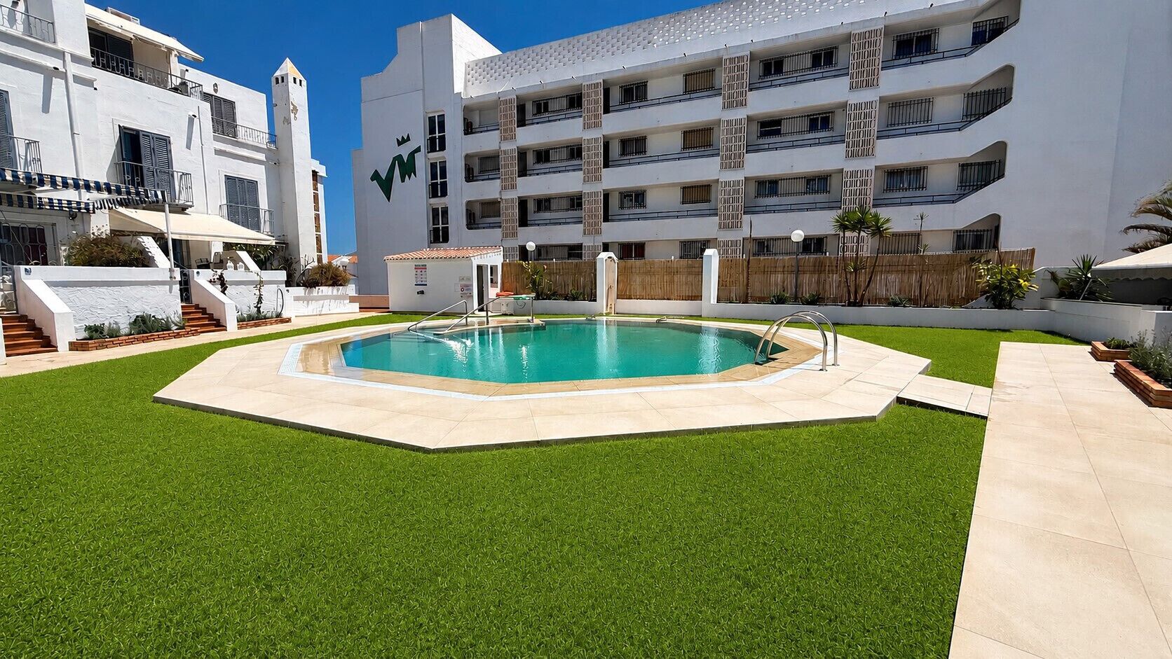 A swimming pool surrounded by artificial grass, with a white building in the background.