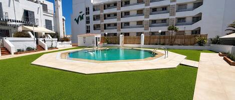 A swimming pool surrounded by artificial grass, with a white building in the background.