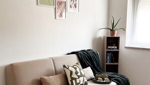 A living room with a beige sofa, a small bookshelf, a potted plant, and framed botanical prints on the wall. - Mysa - Your home 5 min from the airport (Isola Sacra)