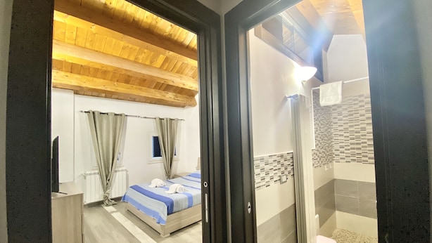 A hotel room with a wooden ceiling, a bed with a blue and white striped bedspread, a window with curtains, a bathroom with a shower and a toilet. - La Dimora Del Golfo (Gaeta)