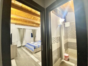 A hotel room with a wooden ceiling, a bed with a blue and white striped bedspread, a window with curtains, a bathroom with a shower and a toilet. - La Dimora Del Golfo (Gaeta)
