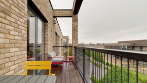 En balkon med trægulv, en gul bænk, en grå sofa og et rødt bord.