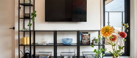 A living room with a black TV stand, shelves with plants and books, and a large window with a view of mountains.