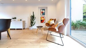 A modern living room with a wooden floor, a black armchair, a brown sofa, a small table, a cactus, and a window with a view of greenery. - Pass the Keys | 49 Byron Square Modern and Spacious 3 Bed Home with Free Parking (Cambridge)